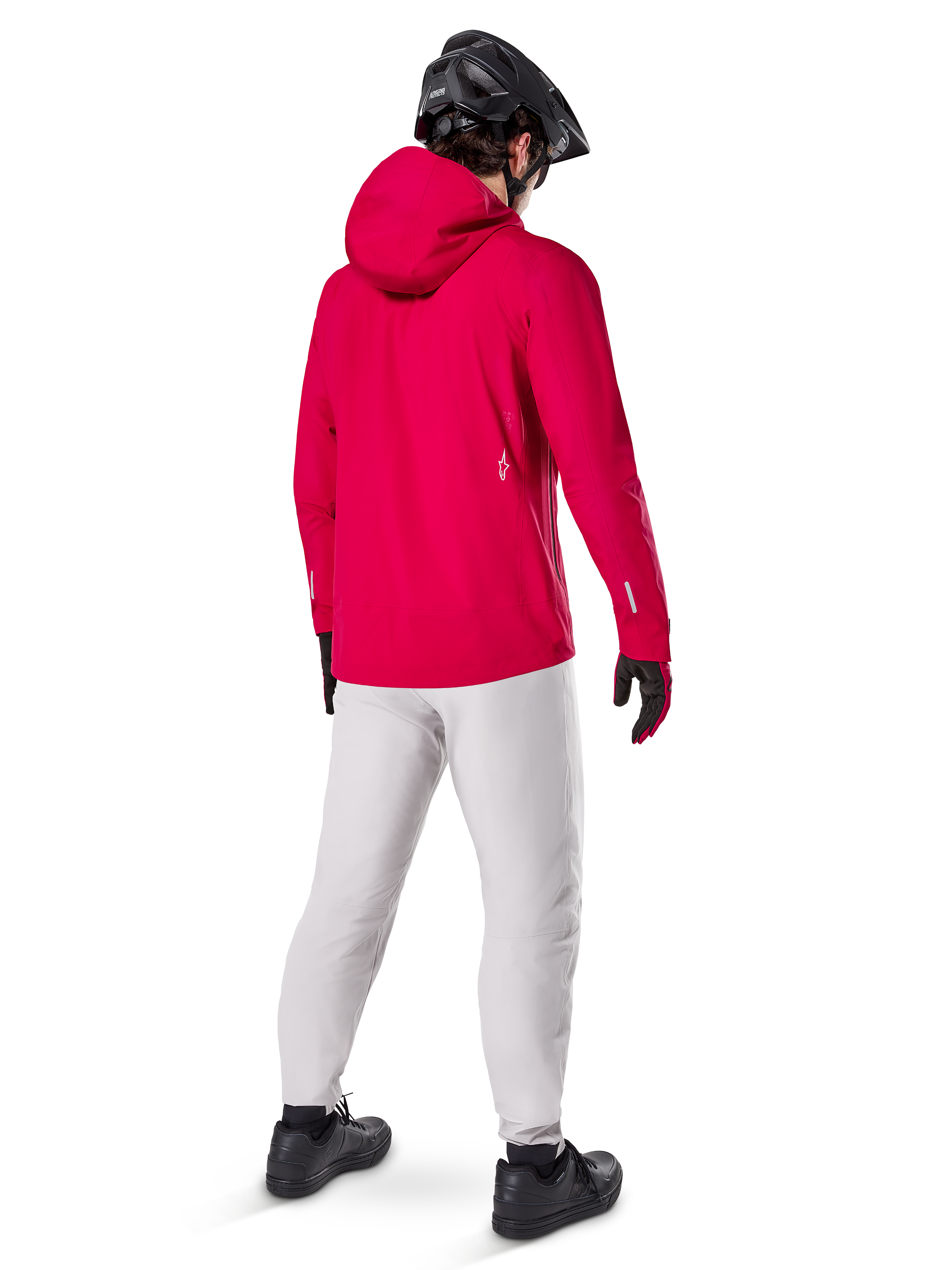 Alpinestars A-Dura Elite vattentät jacka, Red Fluo, sedd snett bakifrån, buren med ljusgrå byxor och svart MTB-hjälm, har huva och reflekterande detaljer, högpresterande cykelregnjacka för stigcykling