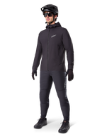 Alpinestars A-Dura Thermal Jacket i svart, bärs av en manlig cyklist med komplett mountainbike-utrustning inklusive svart hjälm, solglasögon och cykelbyxor, lätt isolerad jacka med huva för prestation i kallt väder.