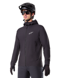 Alpinestars A-Dura Thermal Jacket, svart cykeljacka, design med huva och dragkedja framtill, vit stjärnlogotyp på bröstet, bärs med svart mountainbikehjälm och handskar, sportig funktionsutrustning för terrängcykling i kallt väder