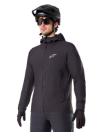 Alpinestars A-Dura Thermal Jacket, svart cykeljacka, design med huva och dragkedja framtill, vit stjärnlogotyp på bröstet, bärs med svart mountainbikehjälm och handskar, sportig funktionsutrustning för terrängcykling i kallt väder