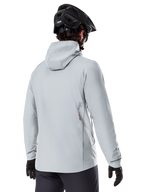 Alpinestars A-Dura Thermal Jacka, Titaniumgrå, vy bakifrån av en cyklist som bär den isolerade jackan med huva, kombinerad med en svart MTB-hjälm och svarta handskar, med en liten Alpinestars-logotyp på nedre delen av ryggen, designad för mountainbiking i kallt väder.