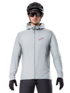 Alpinestars A-Dura Thermal Jacka i Titanium, ljusgrå cykeljacka med huva, röd stjärnlogotyp på bröstet, bärs av en cyklist med svart MTB-hjälm, solglasögon och handskar, isolerad utrustning för mountainbike