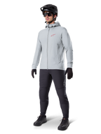 Alpinestars A-Dura Thermal Jacka i Titanium, ljusgrå cykeljacka med huva och en röd Alpinestars-logotyp på bröstet, bärs av en man med svart MTB-hjälm, solglasögon, mörkgrå byxor och svarta handskar, professionell mountainbike-utrustning.