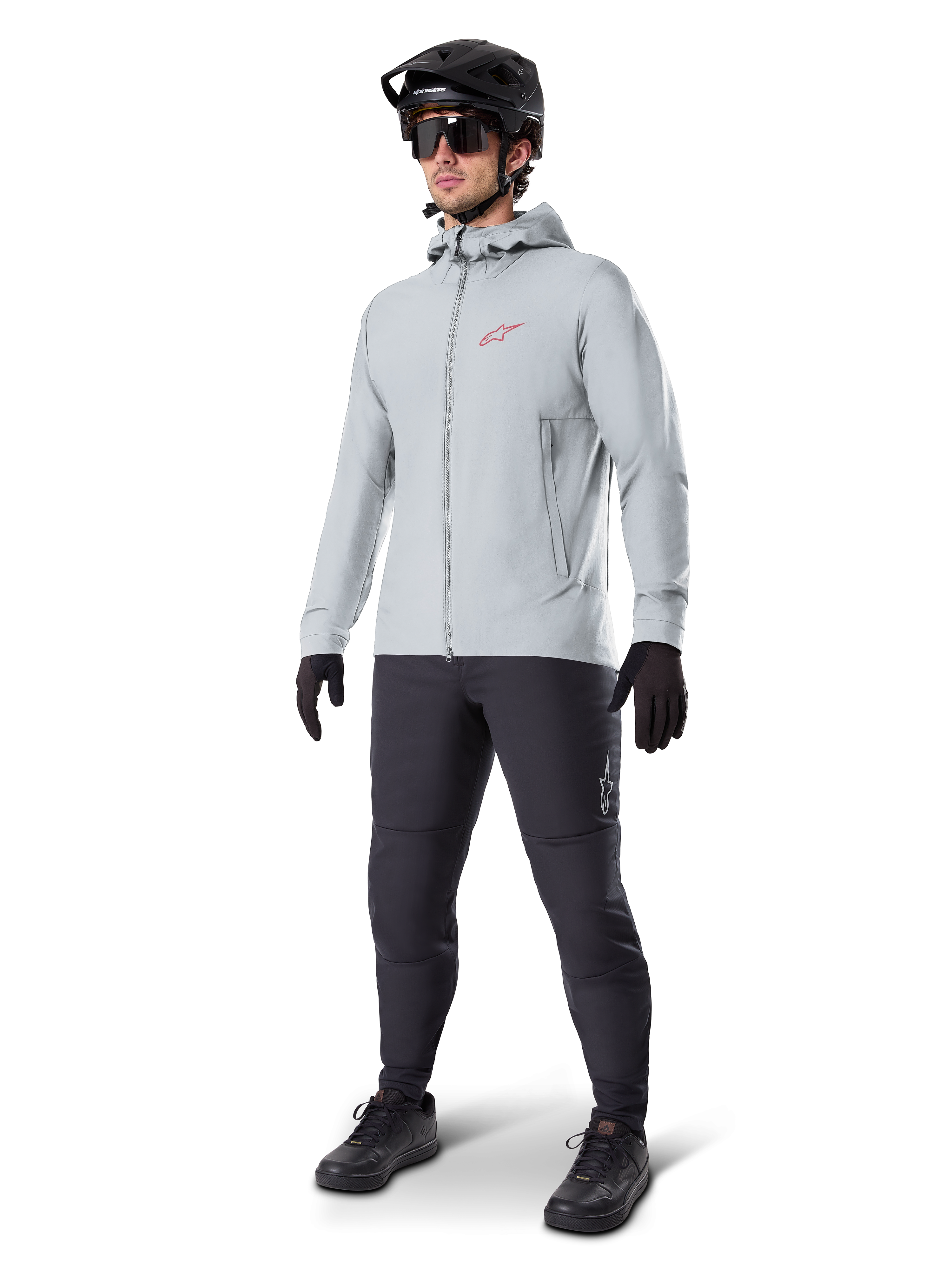 Alpinestars A-Dura Thermal Jacka i Titanium, ljusgrå cykeljacka med huva och en röd Alpinestars-logotyp på bröstet, bärs av en man med svart MTB-hjälm, solglasögon, mörkgrå byxor och svarta handskar, professionell mountainbike-utrustning.
