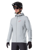 Alpinestars A-Dura Thermal Jacket, ljusgrå isolerad cykeljacka, design med huva och en liten röd logotyp, bärs med svart hjälm och handskar, tekniska mountainbikekläder för cykling i kallt väder