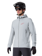 Alpinestars A-Dura Thermal Jacket, ljusgrå isolerad cykeljacka, design med huva och en liten röd logotyp, bärs med svart hjälm och handskar, tekniska mountainbikekläder för cykling i kallt väder