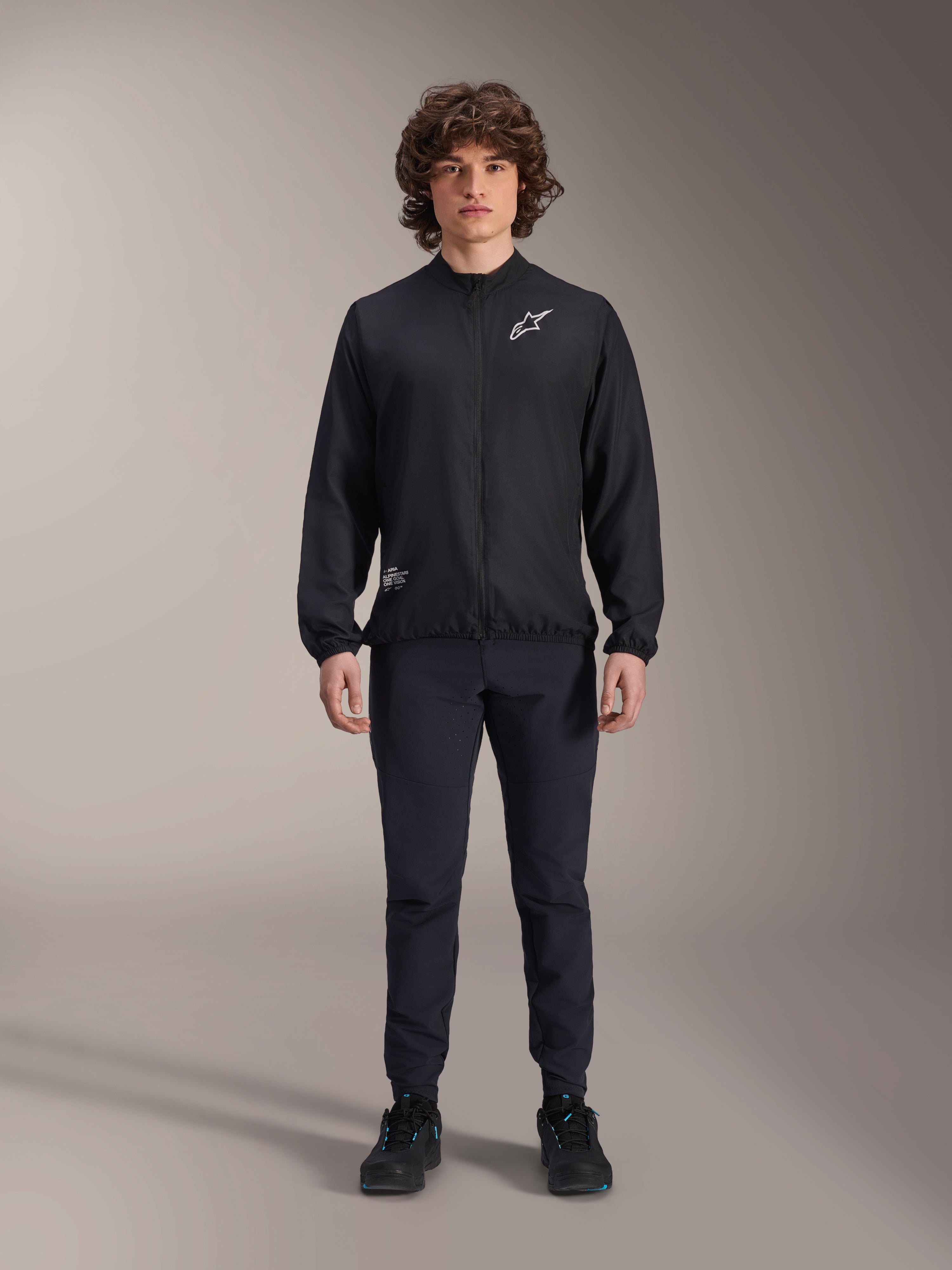 Modell iklädd Alpinestars A-Aria vindjacka i svart, med hel dragkedja fram, hög krage och vit Astars-logotyp på bröstet, matchad med svarta tekniska byxor och svarta skor, prestationsinriktade cykelkläder för mountainbike och utomhusbruk.