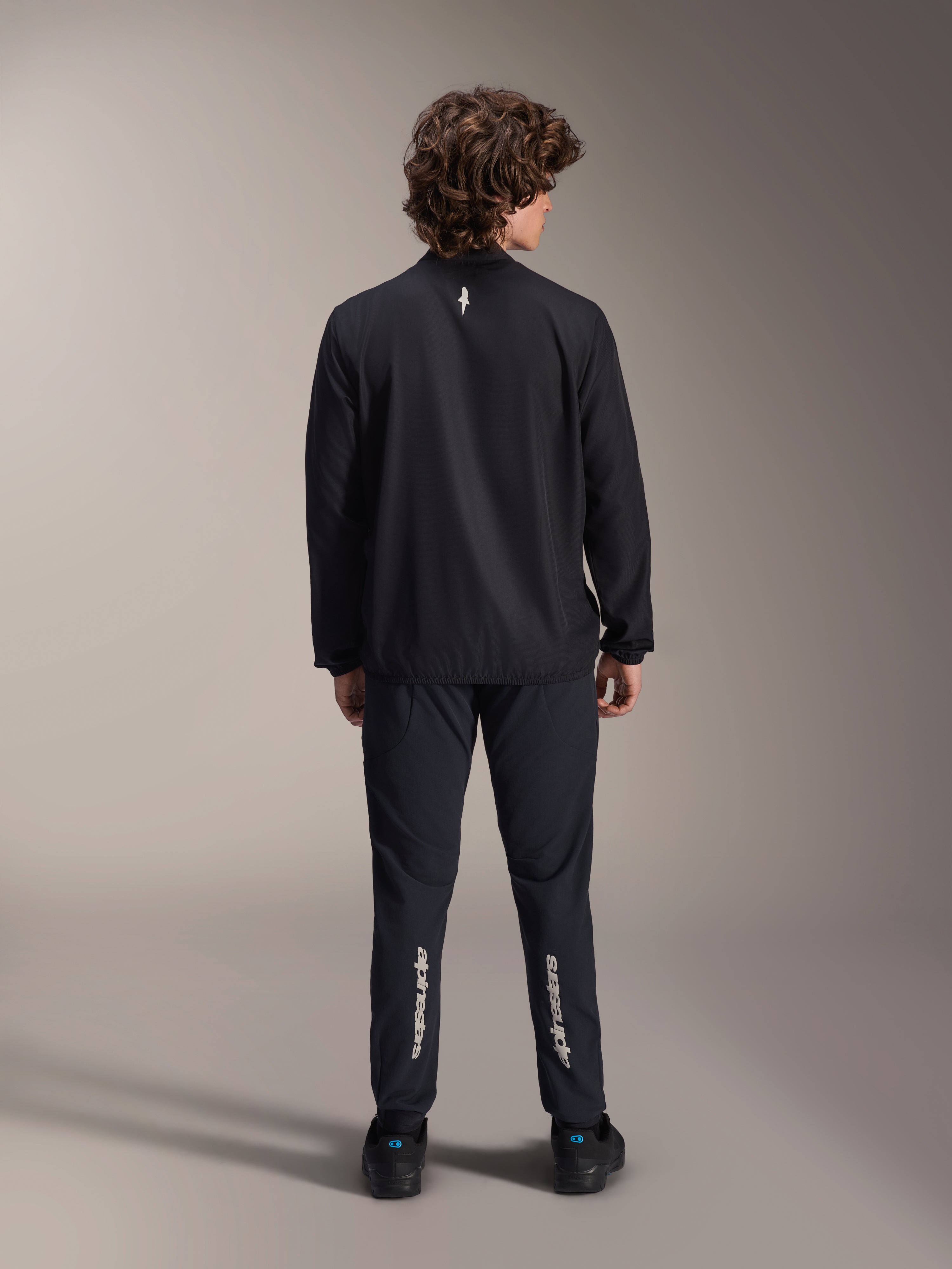 Alpinestars A-Aria vindjacka, svart, vy bakifrån, lätt vindavvisande cykeljacka med en liten vit logotyp på övre ryggen, bärs med matchande svarta byxor med vertikal Alpinestars-branding på vaderna, prestationsinriktad mountainbike-utrustning