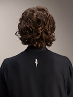 Alpinestars A-Aria vindjacka, svart, närbild bakifrån på axlar och krage, en liten vit logotyp mitt på ryggen, bärs av modell med vågigt brunt hår, minimalistisk cykelutrustning med hög prestanda