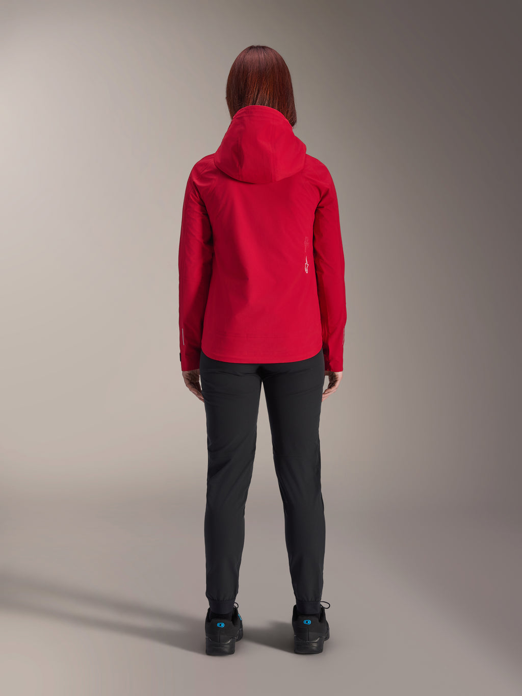 Ryggvy av en kvinna som bär Alpinestars Stella A-Dura Elite Waterproof Jacket i Red Fluo, stylad med svarta byxor och skor, med en skyddande huva och längre bakfåll för mountainbike.