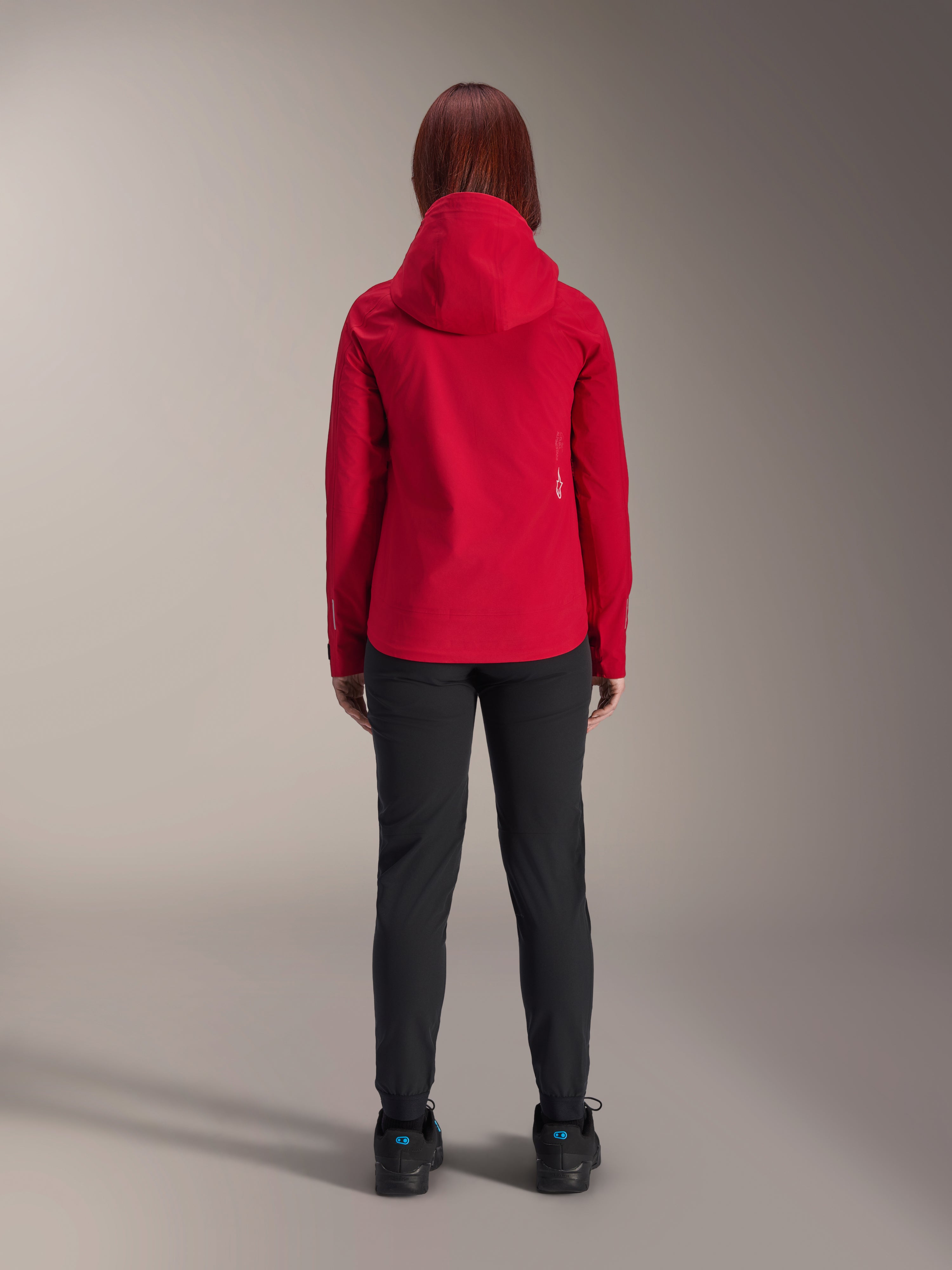 Ryggvy av en kvinna som bär Alpinestars Stella A-Dura Elite Waterproof Jacket i Red Fluo, stylad med svarta byxor och skor, med en skyddande huva och längre bakfåll för mountainbike.
