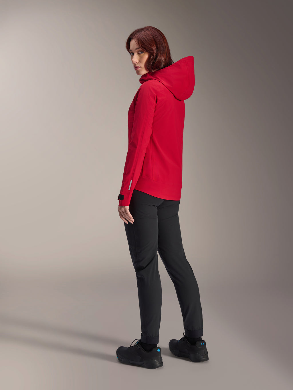 Kvinna bär Alpinestars Stella A-Dura Elite Waterproof Jacket i Red Fluo, kombinerad med svarta tekniska cykelbyxor och svarta skor, trekvartsvy bakifrån som visar den justerbara huvan och den förlängda bakfållen, tekniska mountainbike-kläder.