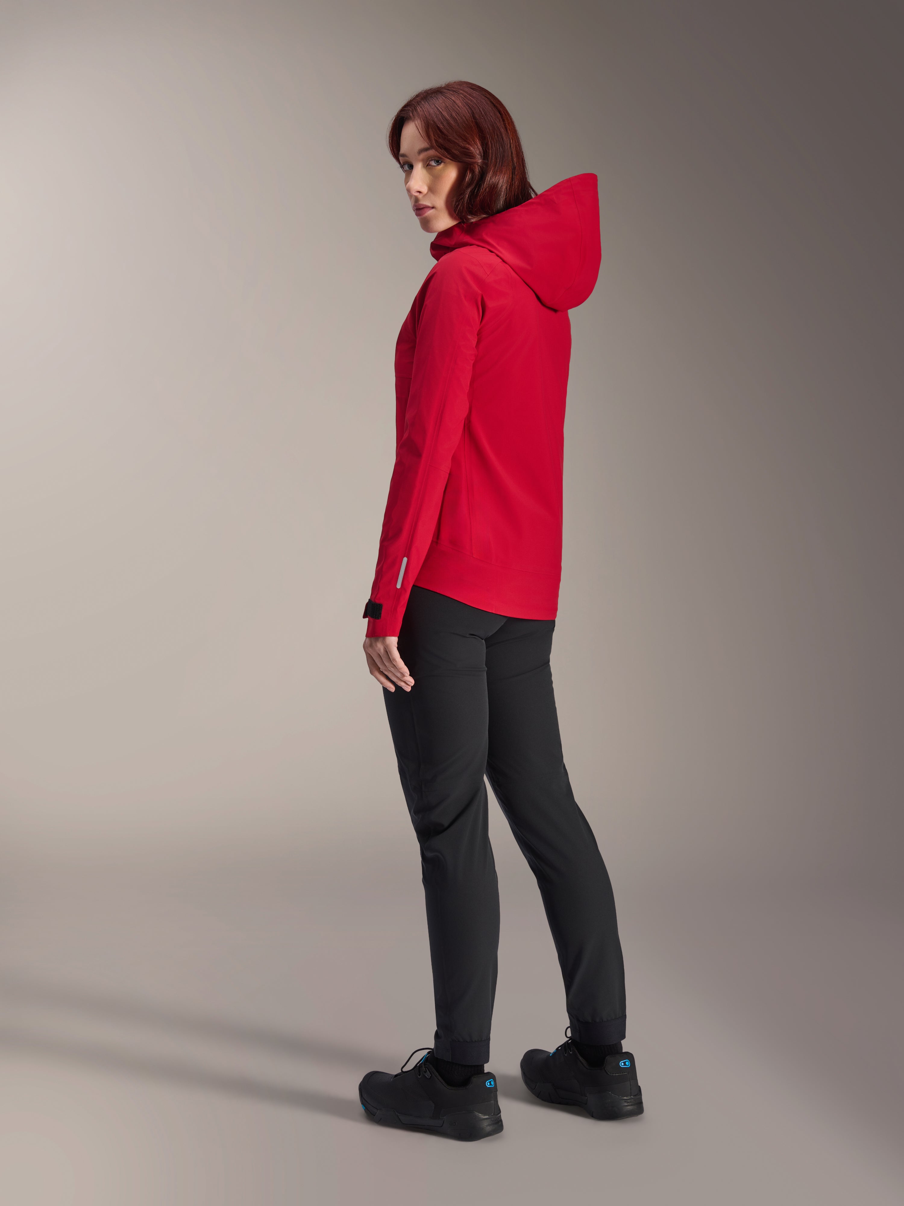 Kvinna bär Alpinestars Stella A-Dura Elite Waterproof Jacket i Red Fluo, kombinerad med svarta tekniska cykelbyxor och svarta skor, trekvartsvy bakifrån som visar den justerbara huvan och den förlängda bakfållen, tekniska mountainbike-kläder.