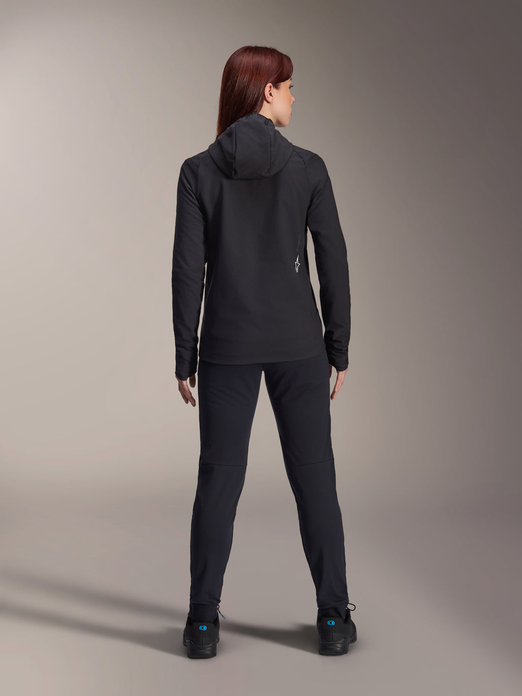 Kvinna som bär Alpinestars Stella A-Dura Thermal Jacket, sedd bakifrån, svart isolerad cykeljacka med huva, matchande svarta byxor och skor, med en liten vit logotyp på nedre delen av ryggen