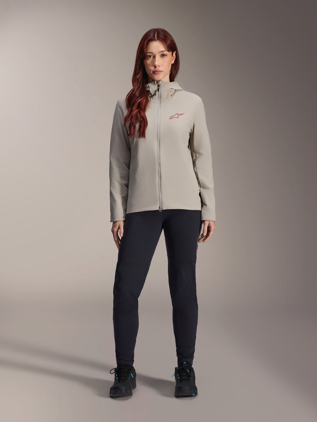 Alpinestars Stella A-Dura Thermal Jacket för dam, mountainbikejacka, titaniumgrå, bärs med svarta tekniska byxor och svarta skor, med en fleecefodrad huva och röd logotyp på bröstet, designad för cykling i kallt väder