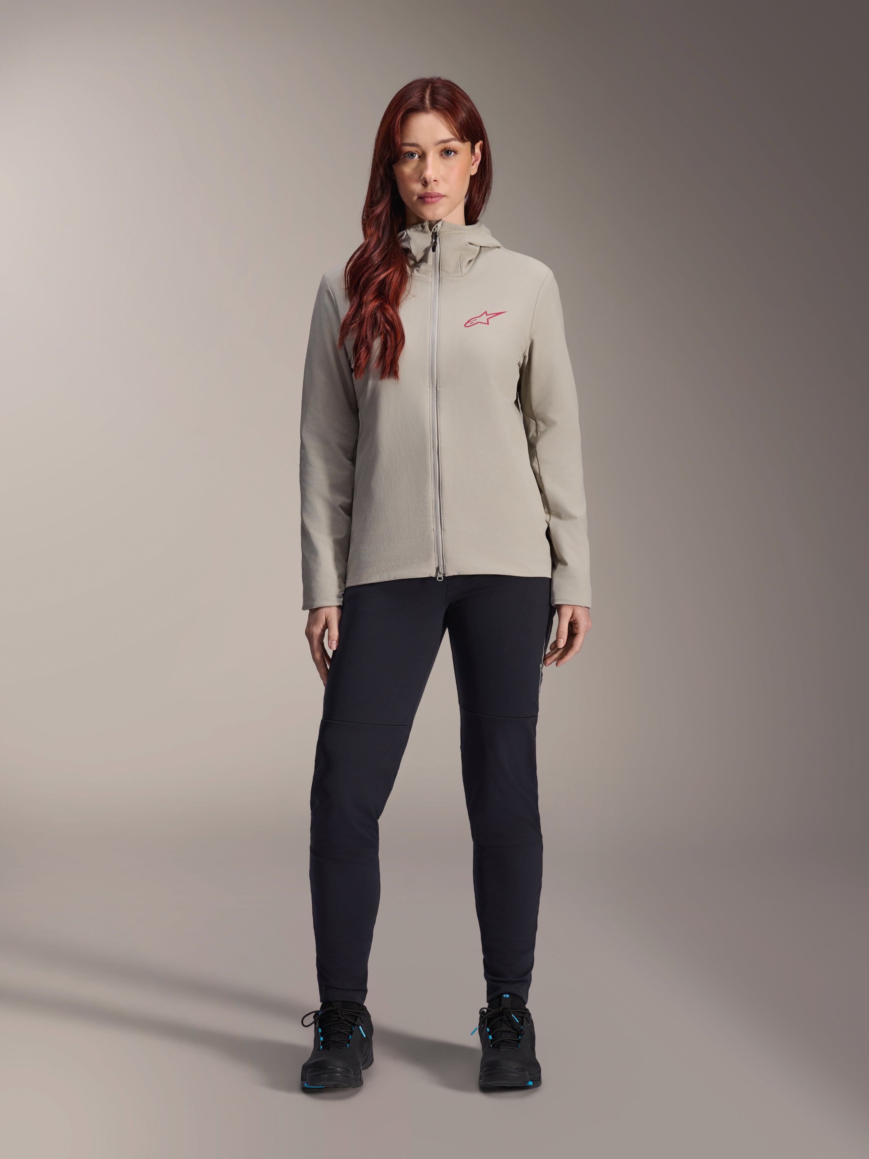 Alpinestars Stella A-Dura Thermal Jacket för dam, mountainbikejacka, titaniumgrå, bärs med svarta tekniska byxor och svarta skor, med en fleecefodrad huva och röd logotyp på bröstet, designad för cykling i kallt väder