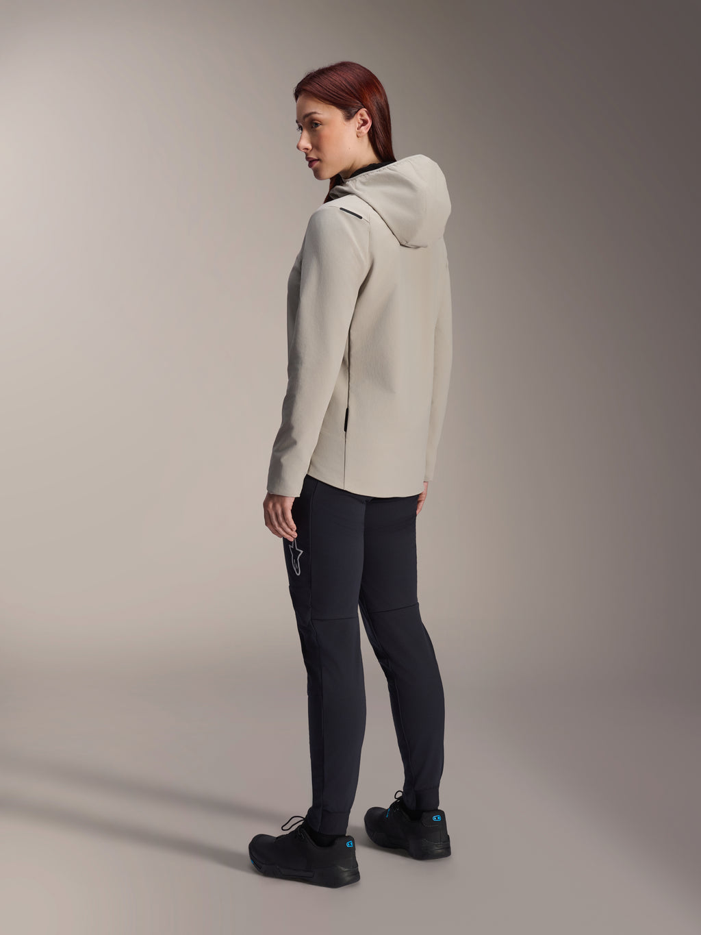 Alpinestars Stella A-Dura Thermal Jacket i titaniumgrått, trekvartsvy bakifrån av en kvinna som bär den isolerade cykeljackan med huva och svarta MTB-byxor, studiobild som visar normal passform och fickdetaljer.