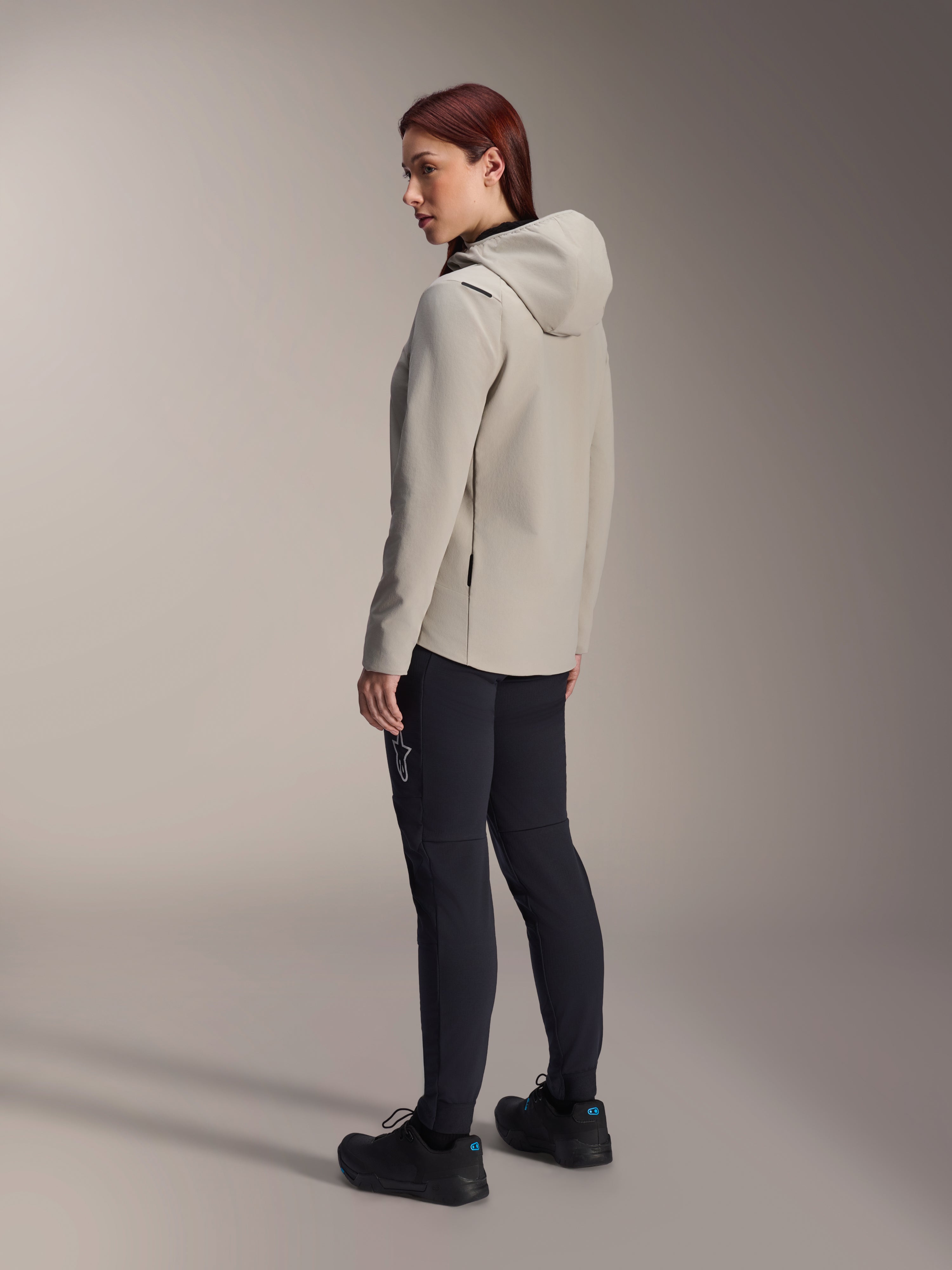 Alpinestars Stella A-Dura Thermal Jacket i titaniumgrått, trekvartsvy bakifrån av en kvinna som bär den isolerade cykeljackan med huva och svarta MTB-byxor, studiobild som visar normal passform och fickdetaljer.