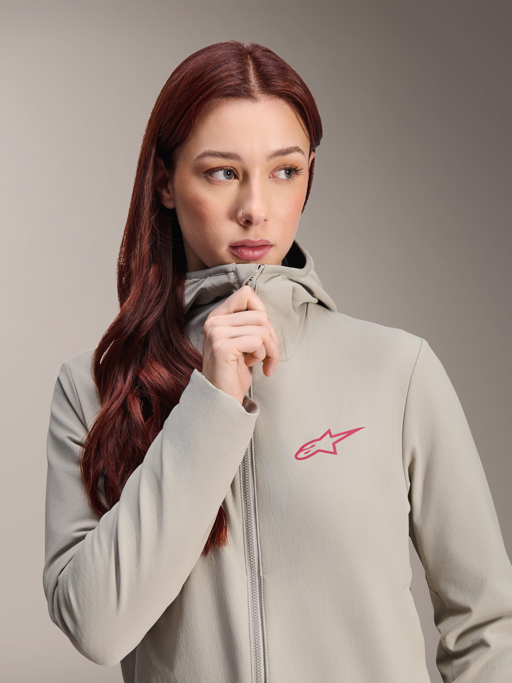 Alpinestars Stella A-Dura Thermal Jacket för dam, titaniumgrå med en rosa Alpinestars-stjärnlogotyp, modell med långt rött hår som bär och drar upp dragkedjan på den huvförsedda jackan, lätt isolerad cykelbeklädnad för kallt väder.