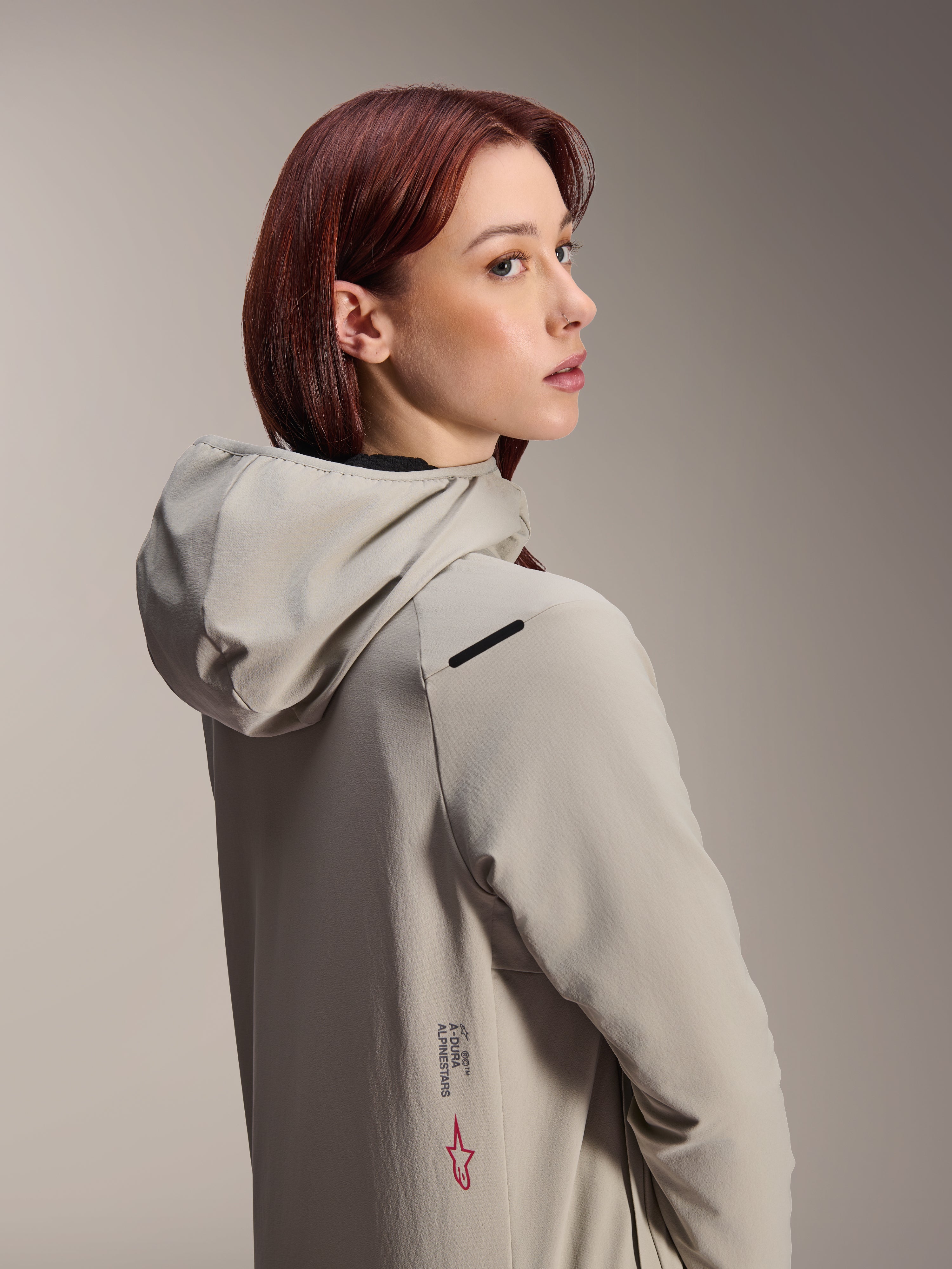Alpinestars Stella A-Dura Thermal Jacket, MTB-jacka för dam, Titaniumgrå, kvinna som ser sig över axeln iklädd en funktionell jacka med huva, med Alpinestars-text och röd stjärnlogotyp på sidan, designad för cykling i kallt väder