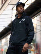 Alpinestars Speeder Chromium Windbreak Jacket, svart vindjacka med huva och vit logotyp på bröstet, bärs av en man med en matchande svart Alpinestars-keps i en urban miljö, avslappnat motorsport-streetwear.