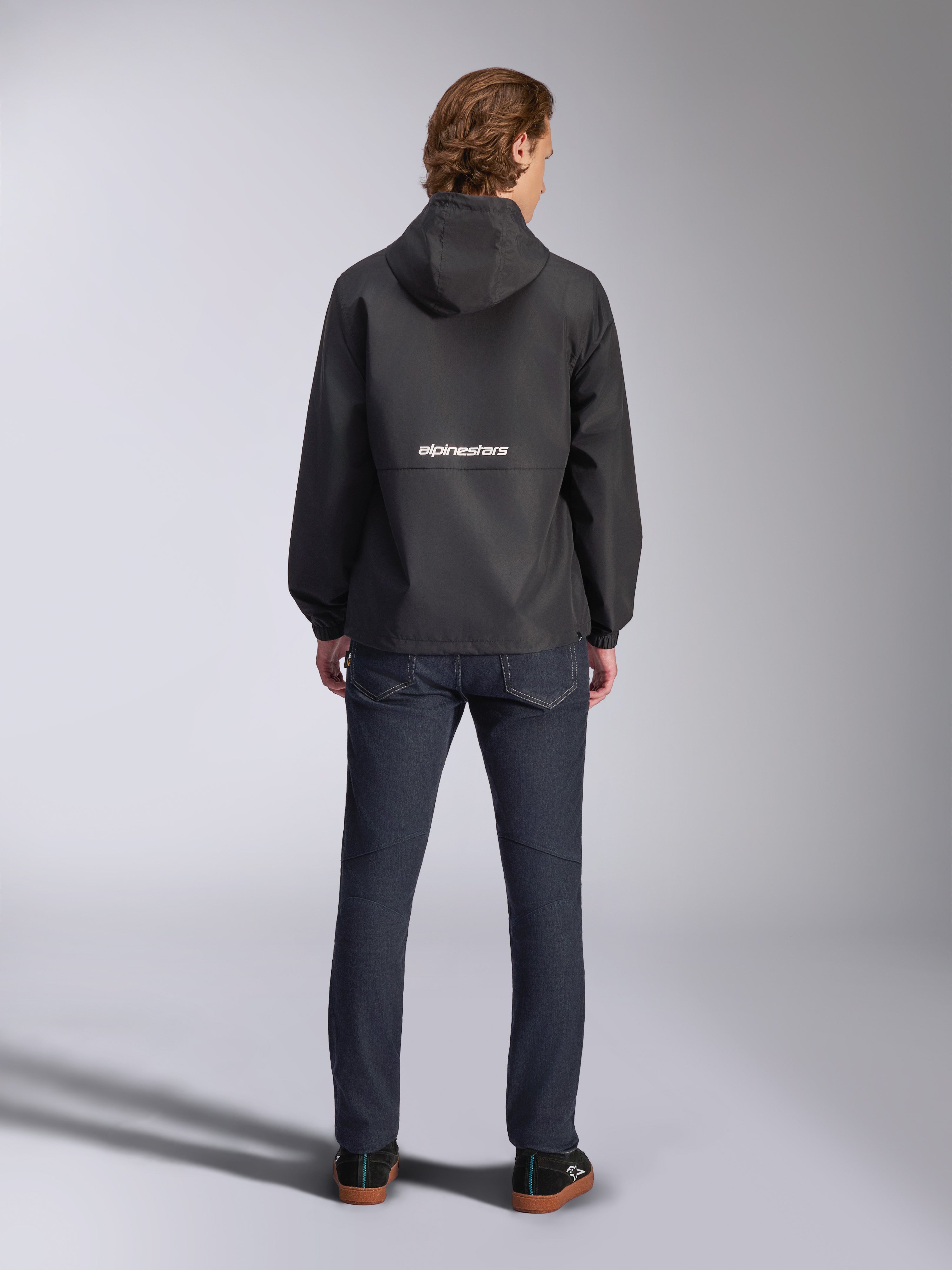 Alpinestars Speeder Chromium vindjacka, svart, sedd bakifrån med vit Alpinestars-logotyp över mitten, modell med huva, bärs av modell med mörka jeans, casual motorsport-inspirerad jacka