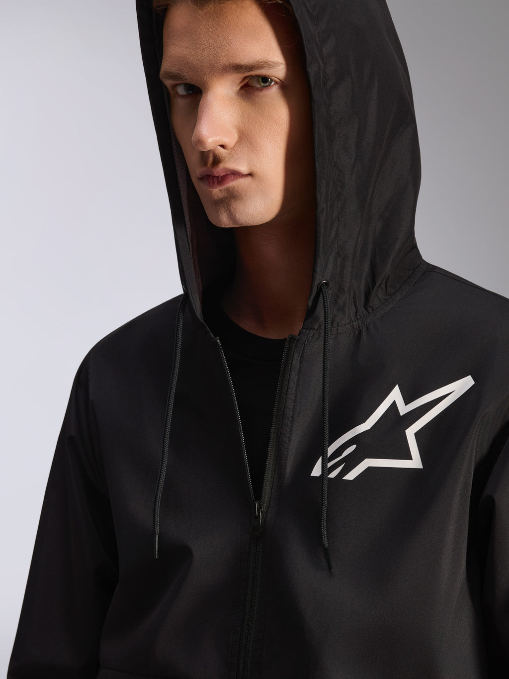 Alpinestars Speeder Chromium vindjacka, svart, närbild av en vindjacka med huva och vit stjärnlogotyp på bröstet, huva med dragsko, lätt textilmaterial, avslappnat motorsportinspirerat streetwear