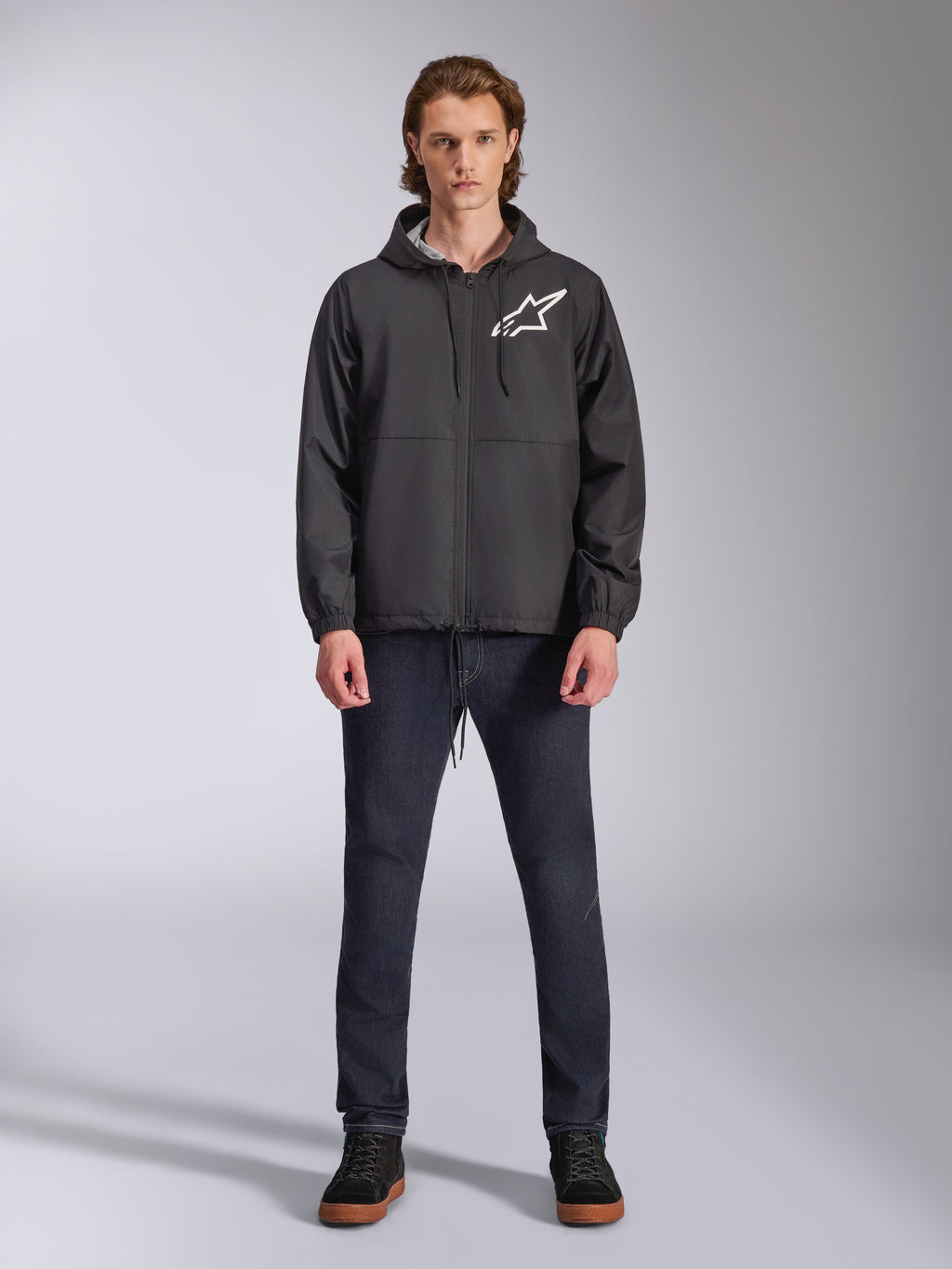 Alpinestars Speeder Chromium vindjacka, svart med vit Astars-logotyp, bärs av modell med mörkblå jeans och svarta sneakers, med huva och dragkedja fram, avslappnad motorsportinspirerad streetwear
