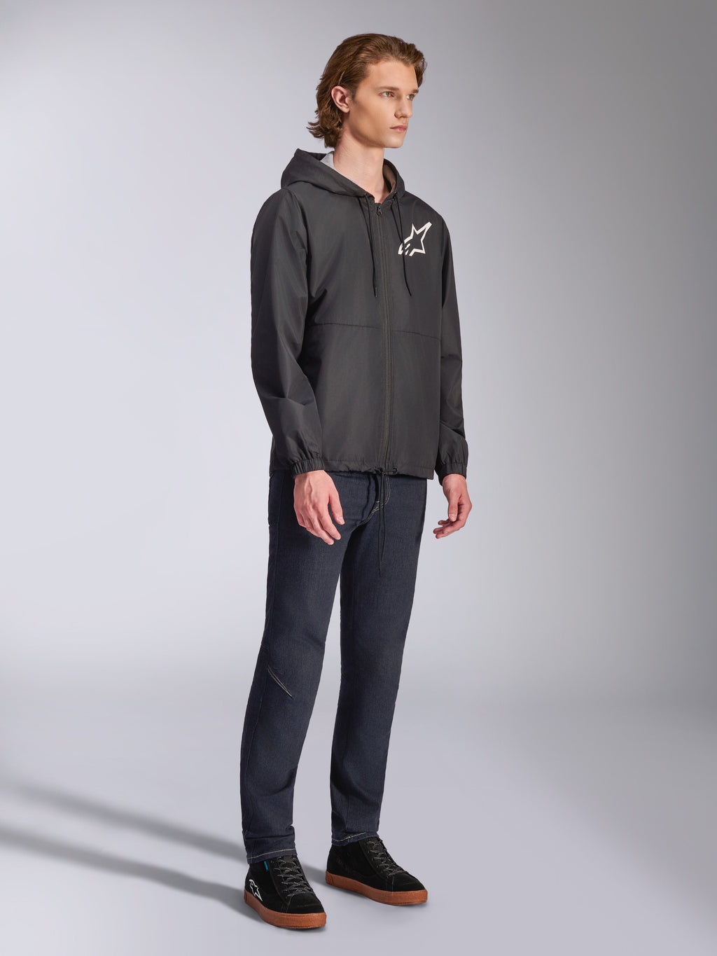 Alpinestars Speeder Chromium Windbreak Jacket, svart, vindjacka med huva och dragkedja fram, vit Alpinestars-stjärnlogotyp på vänster bröst, casual motorsport-streetwear, stylad med mörka byxor och svarta sneakers i studiomiljö.