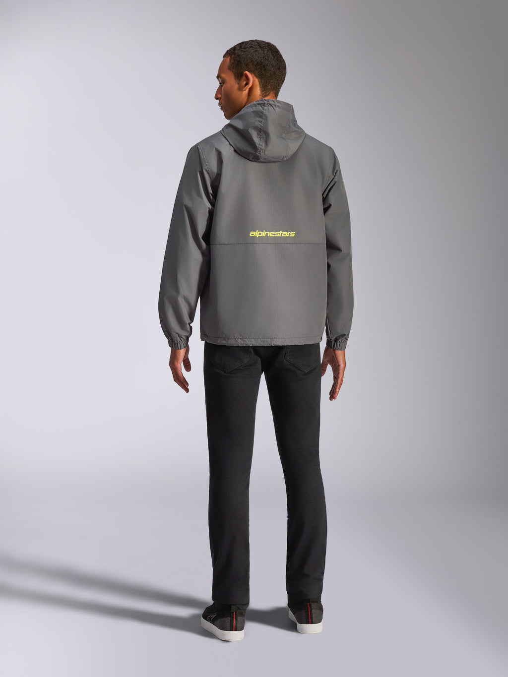 Alpinestars Speeder Chromium vindjacka, mörkgrå och neongul, vy bakifrån som visar en gul Alpinestars-logotyp över mitten, lätt vindjacka med huva och elastiska muddar, avslappnat motorsportinspirerat streetwear