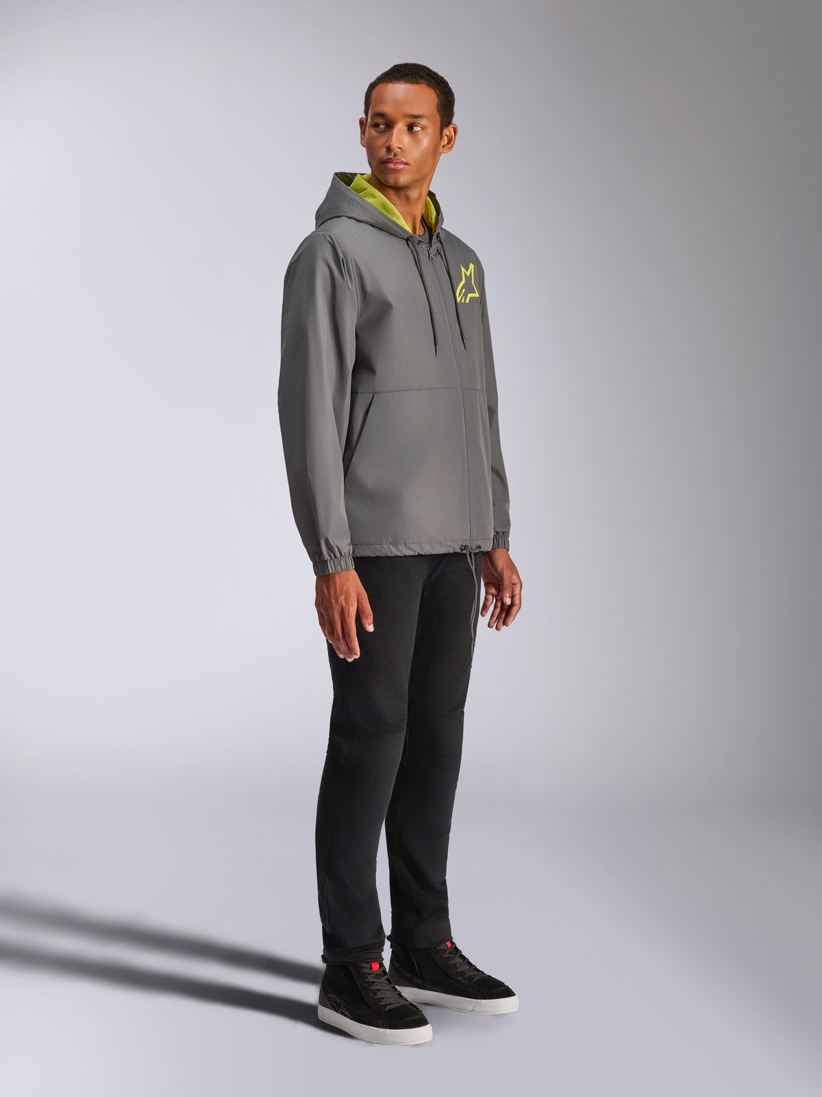 Alpinestars Speeder Chromium vindjacka, antracitgrå med neongula detaljer, manlig modell i vindjacka med huva, gul stjärnlogotyp på bröstet, matchad med svarta byxor och svarta sneakers, avslappnad motorsport-inspirerad streetwear