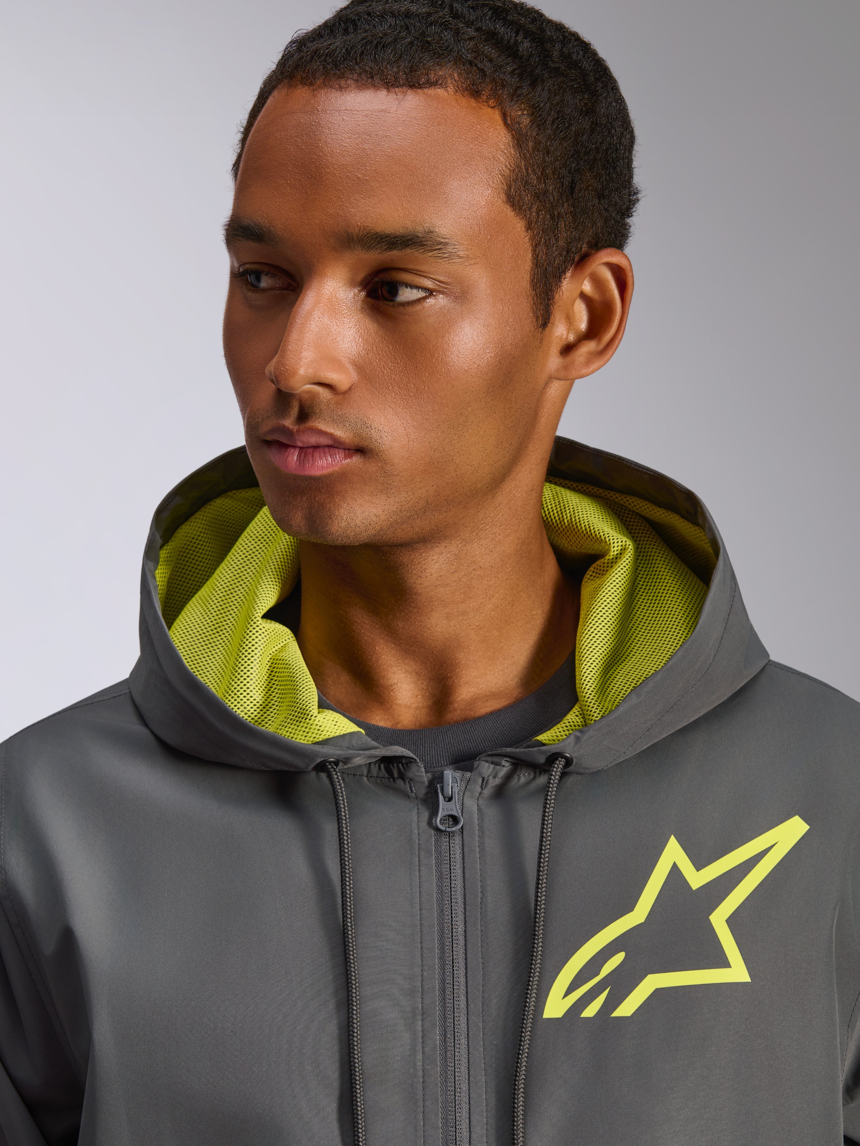 Alpinestars Speeder Chromium vindjacka, mörkgrå med varselgula detaljer, med en gul stjärnlogotyp på bröstet och matchande meshfoder i huvan, lätt och ledig vindjacka