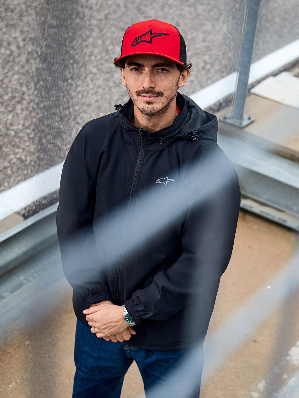 Man som bär Alpinestars Overhaul Racer Jacket i svart, en vattenavvisande regnskaljacka med huva och en liten logotyp på bröstet, matchad med en röd Alpinestars-keps, casual motorsport-lifestylekläder