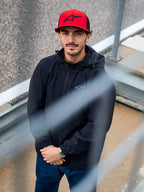 Man som bär Alpinestars Overhaul Racer Jacket i svart, en vattenavvisande regnskaljacka med huva och en liten logotyp på bröstet, matchad med en röd Alpinestars-keps, casual motorsport-lifestylekläder