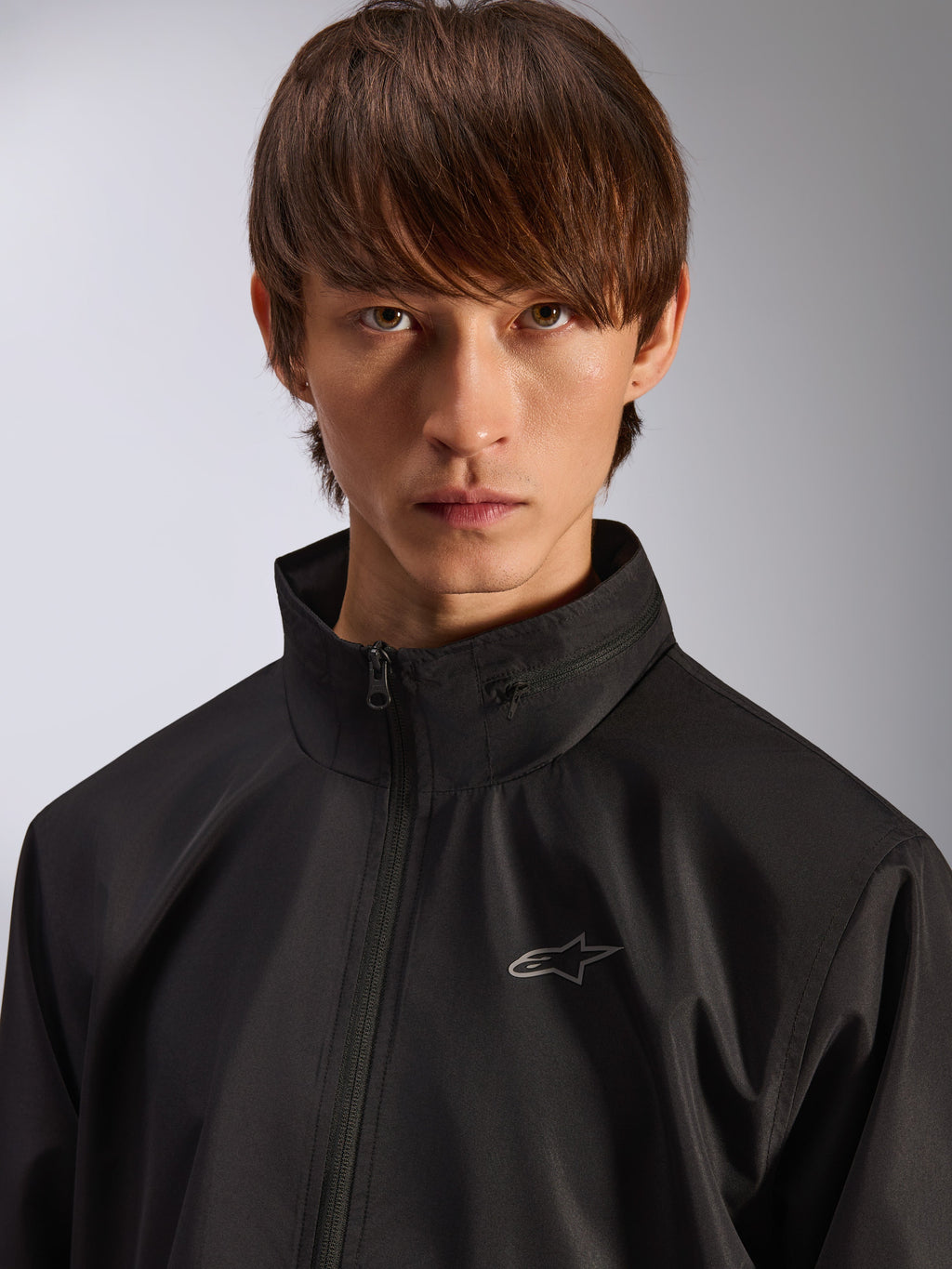 Alpinestars Overhaul Racer Jacket, svart regnskaljacka, närbildsporträtt av en manlig modell som bär jackan, med hög krage och liten silverfärgad Astars-logotyp på bröstet, motorsportinspirerat vardagsplagg