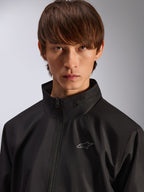 Alpinestars Overhaul Racer Jacket, svart regnskaljacka, närbildsporträtt av en manlig modell som bär jackan, med hög krage och liten silverfärgad Astars-logotyp på bröstet, motorsportinspirerat vardagsplagg