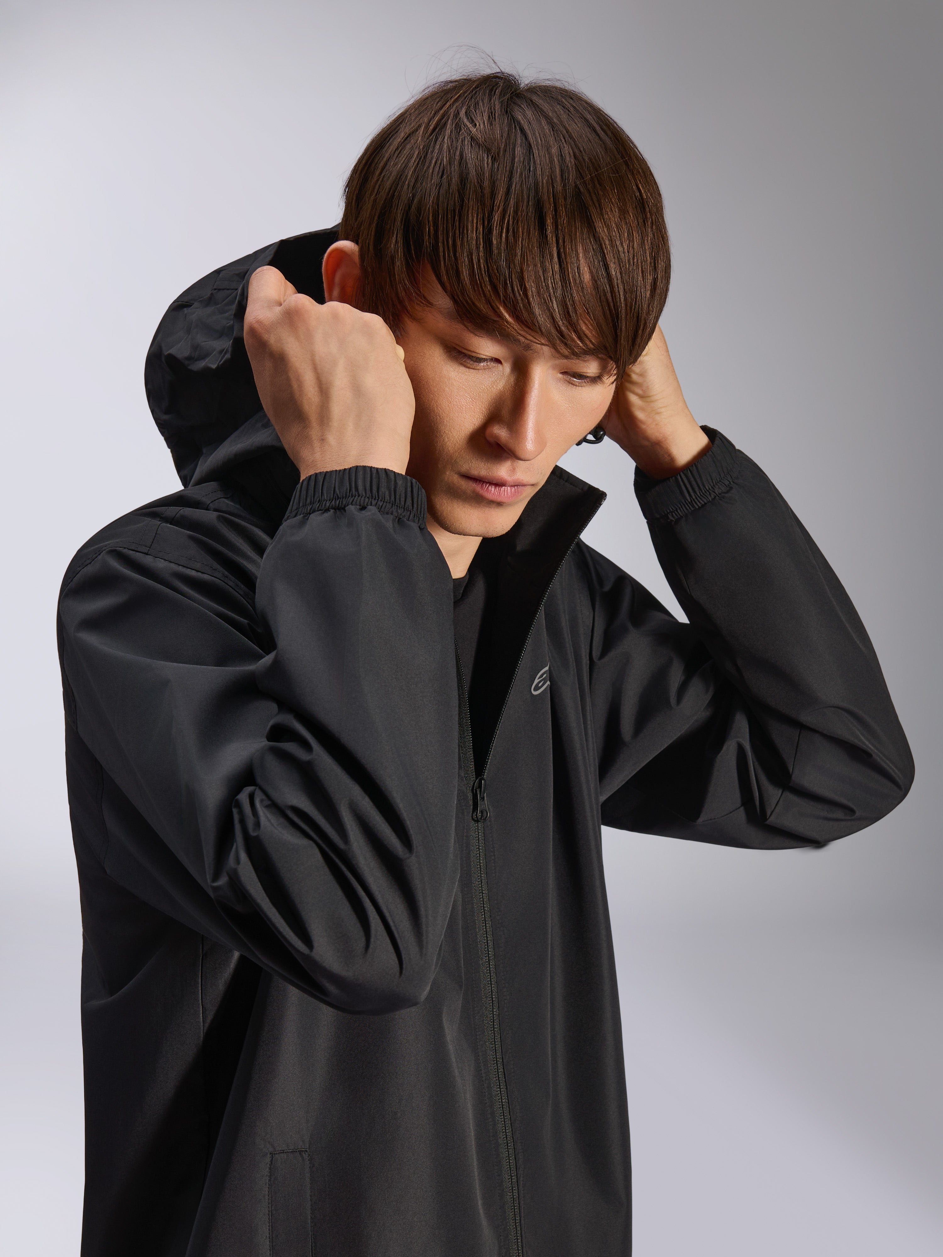 Alpinestars Overhaul Racer Jacket, svart regnskaljacka, bärs av modell med huvan uppfälld, tillverkad i vattenavvisande 2,5L-tyg, elastiska muddar och diskret logotyp på bröstet, vardagligt motorsport-inspirerat ytterplagg