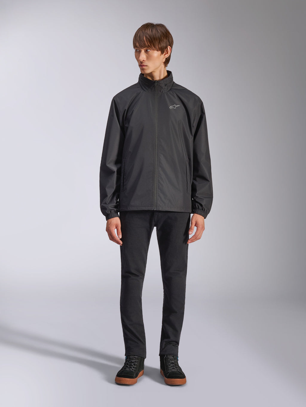 Alpinestars Overhaul Racer Jacket, svart regnskalsjacka för herrar, helfigursvy på en modell kombinerad med svarta byxor och vardagliga sneakers, liten vit logotyp på bröstet, vattenavvisande material och avslappnad motorsportinspirerad design