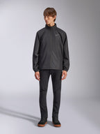 Alpinestars Overhaul Racer Jacket, svart regnskalsjacka för herrar, helfigursvy på en modell kombinerad med svarta byxor och vardagliga sneakers, liten vit logotyp på bröstet, vattenavvisande material och avslappnad motorsportinspirerad design