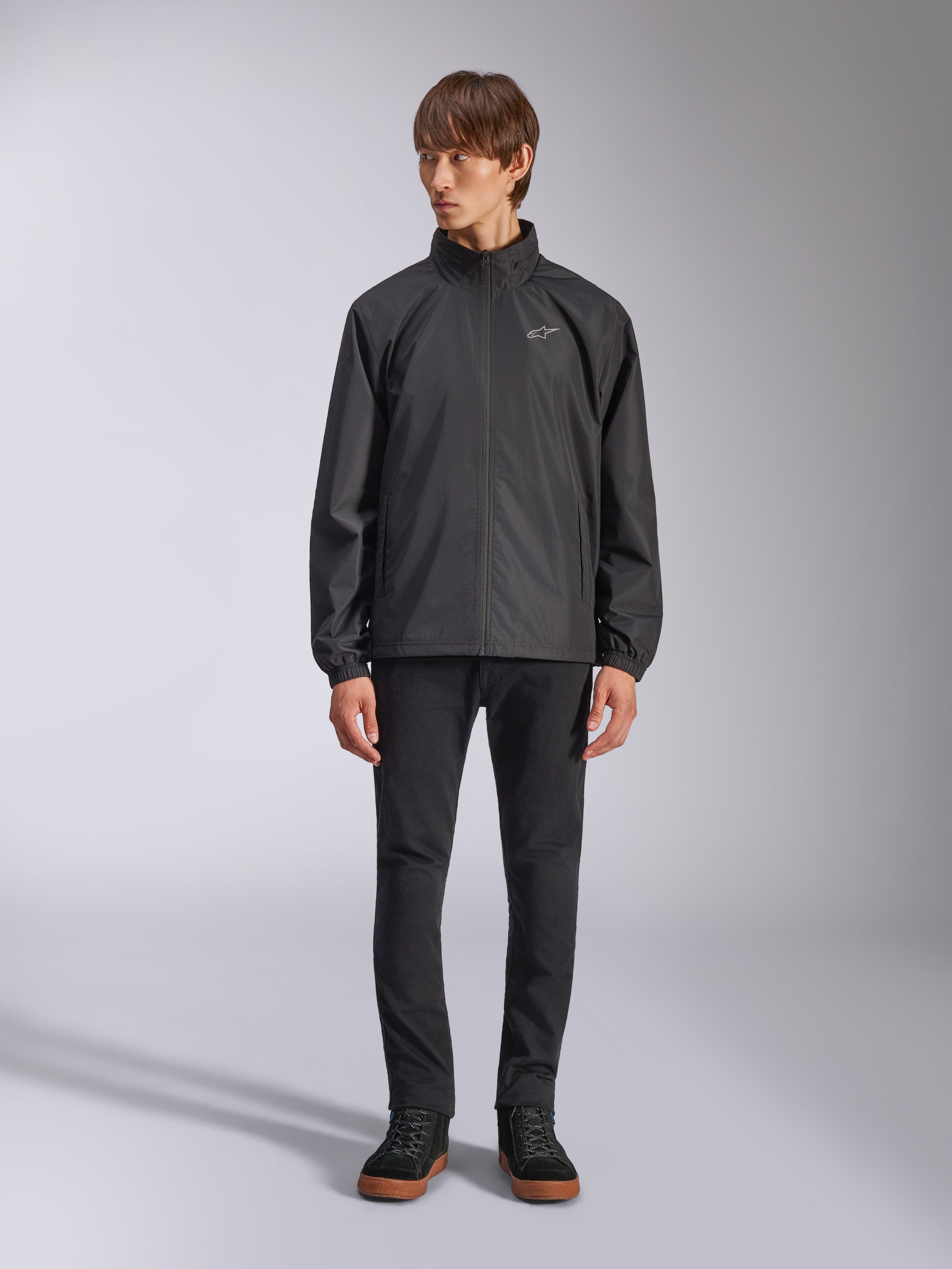 Alpinestars Overhaul Racer Jacket, svart regnskalsjacka för herrar, helfigursvy på en modell kombinerad med svarta byxor och vardagliga sneakers, liten vit logotyp på bröstet, vattenavvisande material och avslappnad motorsportinspirerad design
