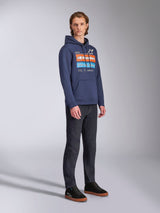 Alpinestars Harken Hoodie in Navy Blue, worn by a male model with dark denim pants and black sneakers, featuring a large chest graphic with orange and light blue accents, Alpinestars wordmark, and kangaroo pocket.