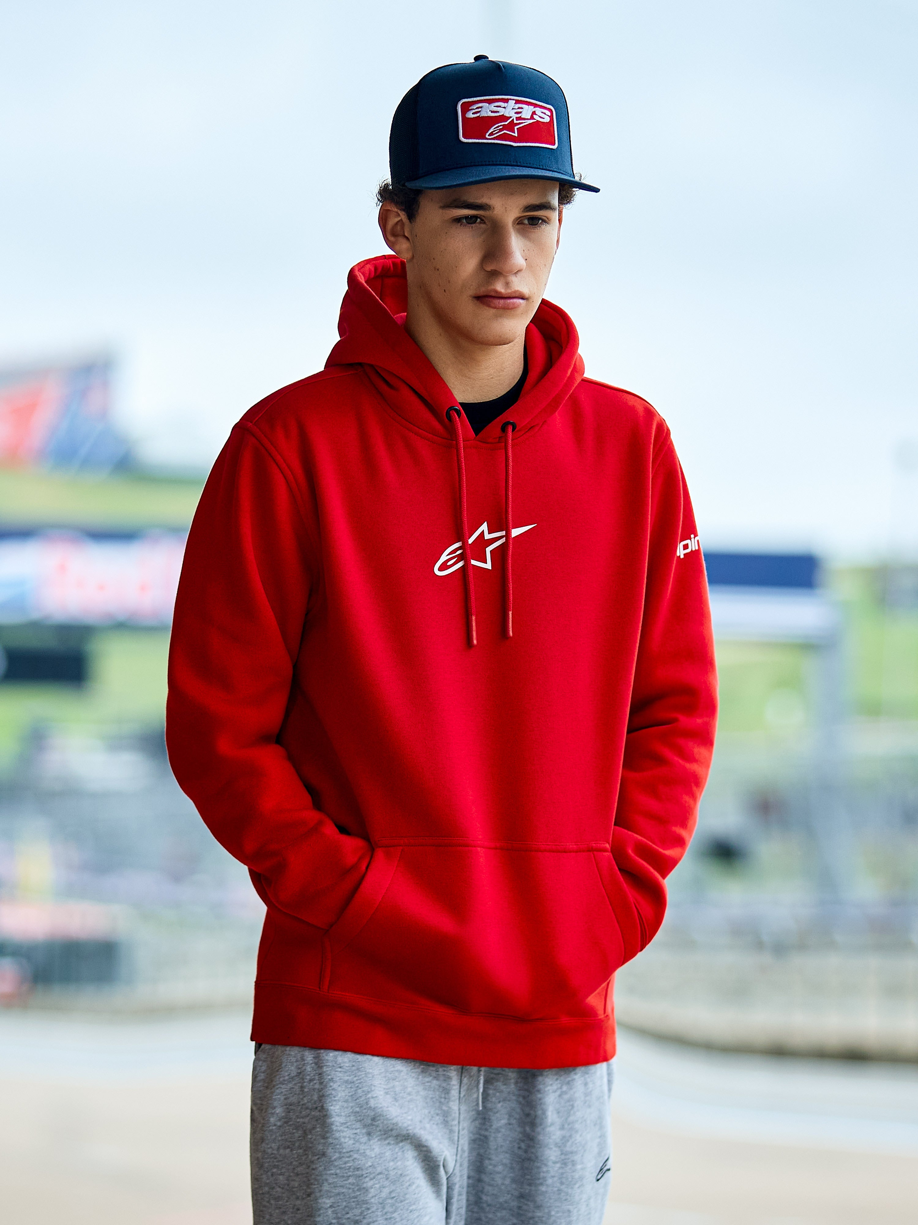 Alpinestars Frontal Hoodie, röd huvtröja i fleece, vit Astars-logotyp på bröstet, känguruficka och huva med dragsko, bärs av en man i blå keps, avslappnat motorsport-streetwear