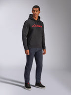 Alpinestars Blaze V3 Hoodie, black pullover fleece featuring a red Alpinestars logo on the chest, worn by a male model with dark denim jeans and black sneakers, casual motorsport-inspired streetwear