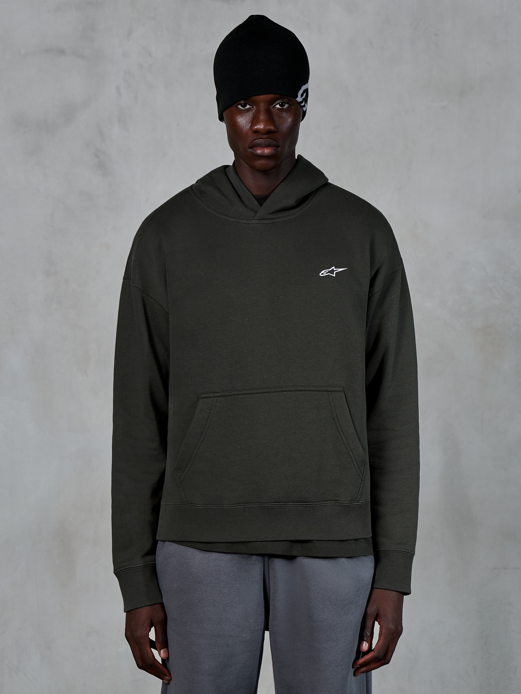 Alpinestars Essentials Ageless Huvtröja, avslappnad streetwear, Off Black pullover med en liten vit Alpinestars-logotyp på bröstet, bärs med en svart mössa, minimalistisk design med känguruficka framtill och plaggstenstvättad finish.