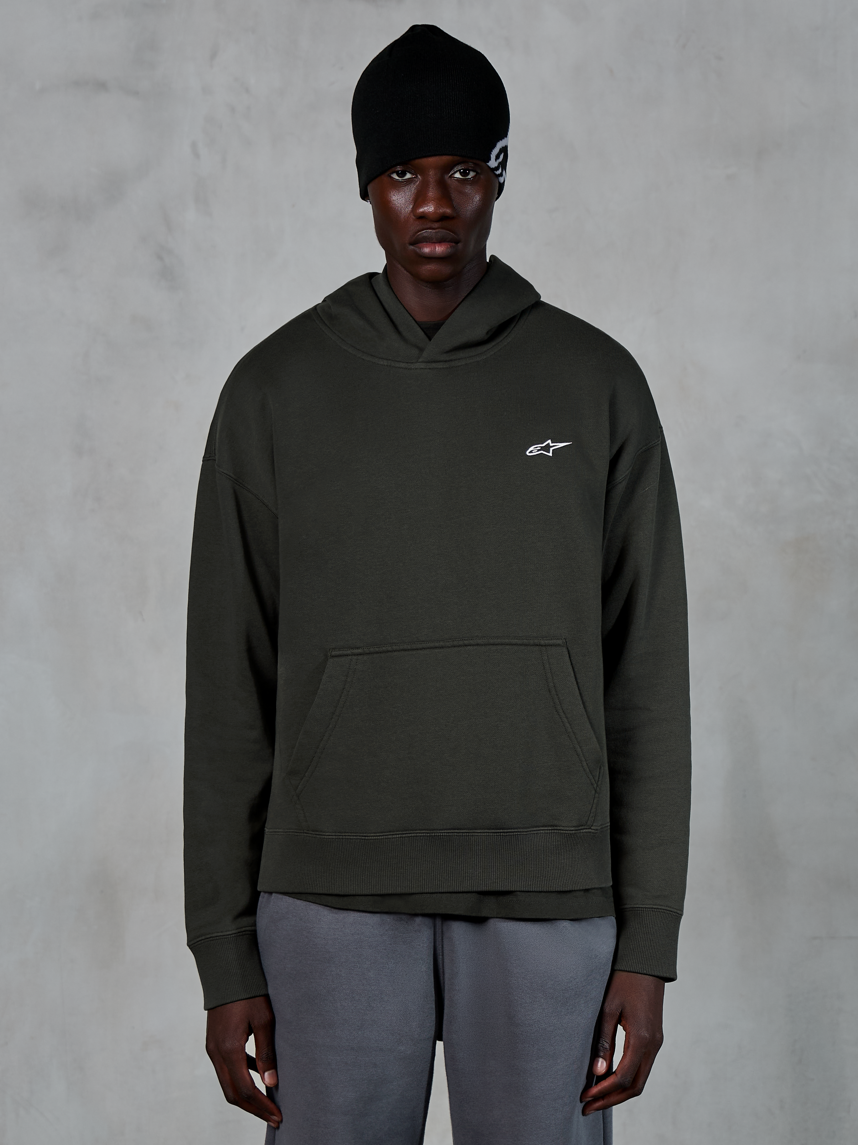 Alpinestars Essentials Ageless Huvtröja, avslappnad streetwear, Off Black pullover med en liten vit Alpinestars-logotyp på bröstet, bärs med en svart mössa, minimalistisk design med känguruficka framtill och plaggstenstvättad finish.
