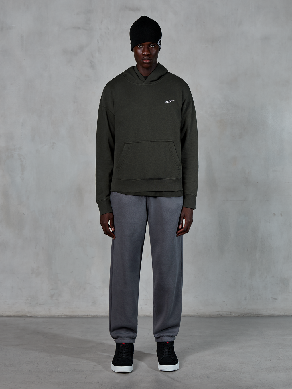 Modell som bär Alpinestars Essentials Ageless Hoodie, casual streetwear, Off Black med liten vit logotyp på bröstet, kombinerad med grå joggers, svart mössa och svarta sneakers, minimalistisk motorsportinspirerad design
