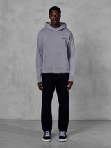 Alpinestars Essentials Ageless Hoodie, silver, manlig modell i avslappnad streetwear, liten svart Alpinestars-logotyp på bröstet, känguruficka framtill, minimalistisk design, matchad med svarta byxor och sneakers.