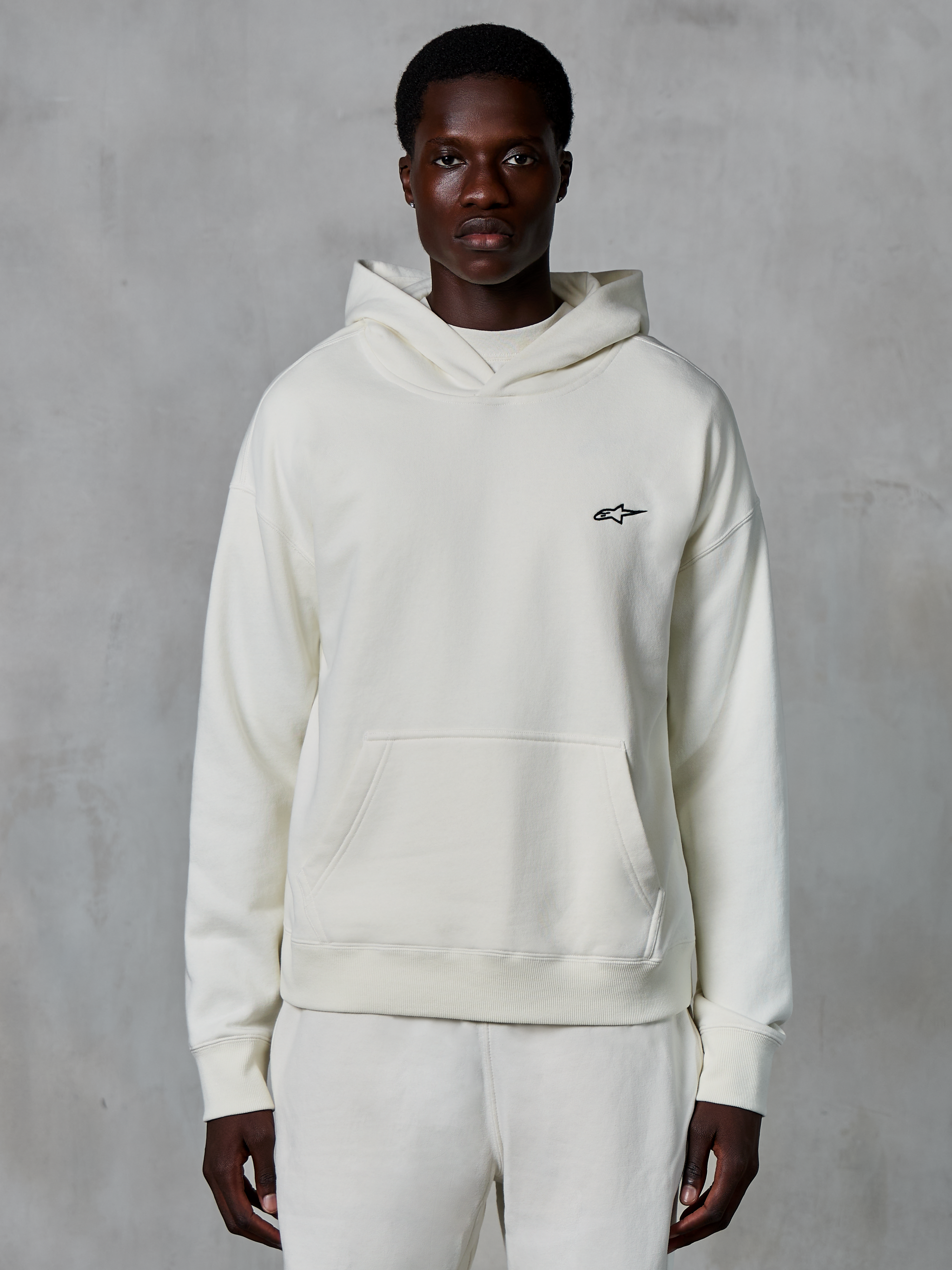 Alpinestars Essentials Ageless Hoodie, off-white, bärs av en manlig modell, liten svart logotyp på bröstet, känguruficka fram, långa ärmar, avslappnat motorsportinspirerat streetwear