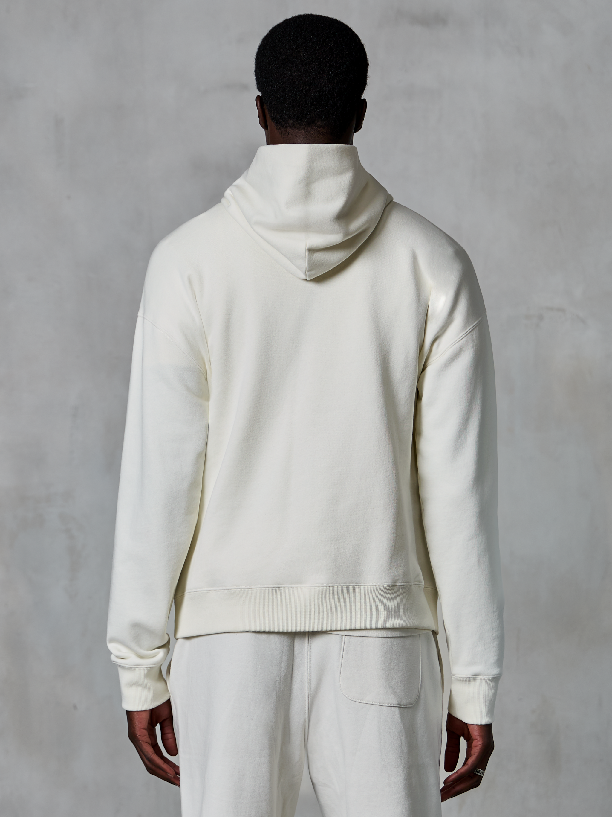 Alpinestars Essentials Ageless Hoodie, off-white pullover-huvtröja, sedd bakifrån på modell, ren minimalistisk design, matchande vardagsbyxor, avslappnad passform för vardagskomfort