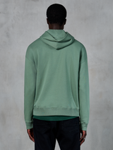 Alpinestars Essentials Ageless Hoodie, avslappnade sportkläder, kaktusgrön, sedd bakifrån på modell, plaggstenstvättad finish, matchad med svarta byxor, minimalistisk livsstilsdesign